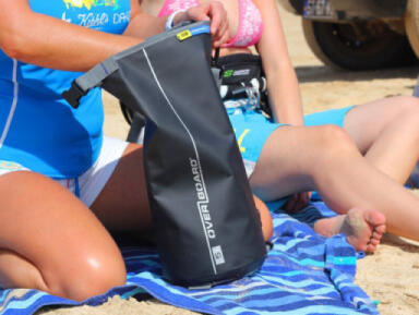 Two people sitting on a beach whilst retrieving personal items from a black 5 litre OverBoard Dry Bag.