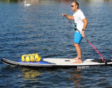 A man on a paddleboard with an OverBoard paddleboard dry bag strapped to the board and an OverBoard phone dry case and lanyard around his neck.
