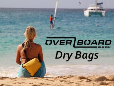 A lady sitting on a sandy beach with her OverBoard Dry Bag on her shoulder with boats on the sea in the distance.